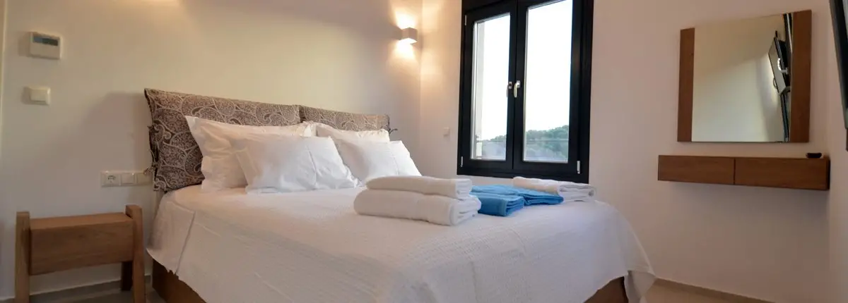 A bedroom with a double bed, a side window, shower towels and a mirror of a private villa at Parga, Greece.