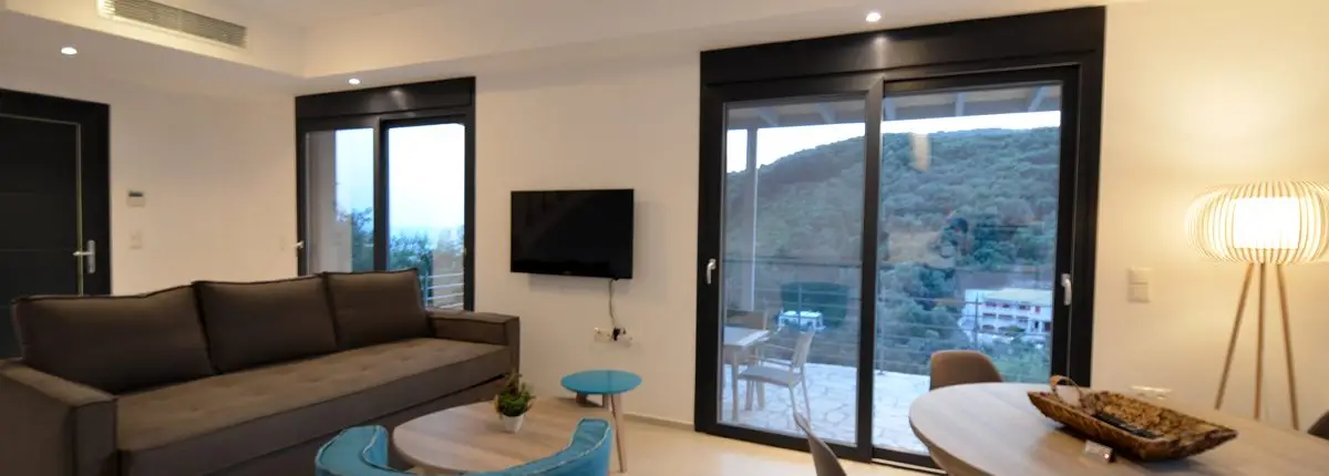 Living room with a sofa, coffee table, dinner table, TV and large balcony doors leading to the balcony at the base floor of a private villa at Parga, Greece.